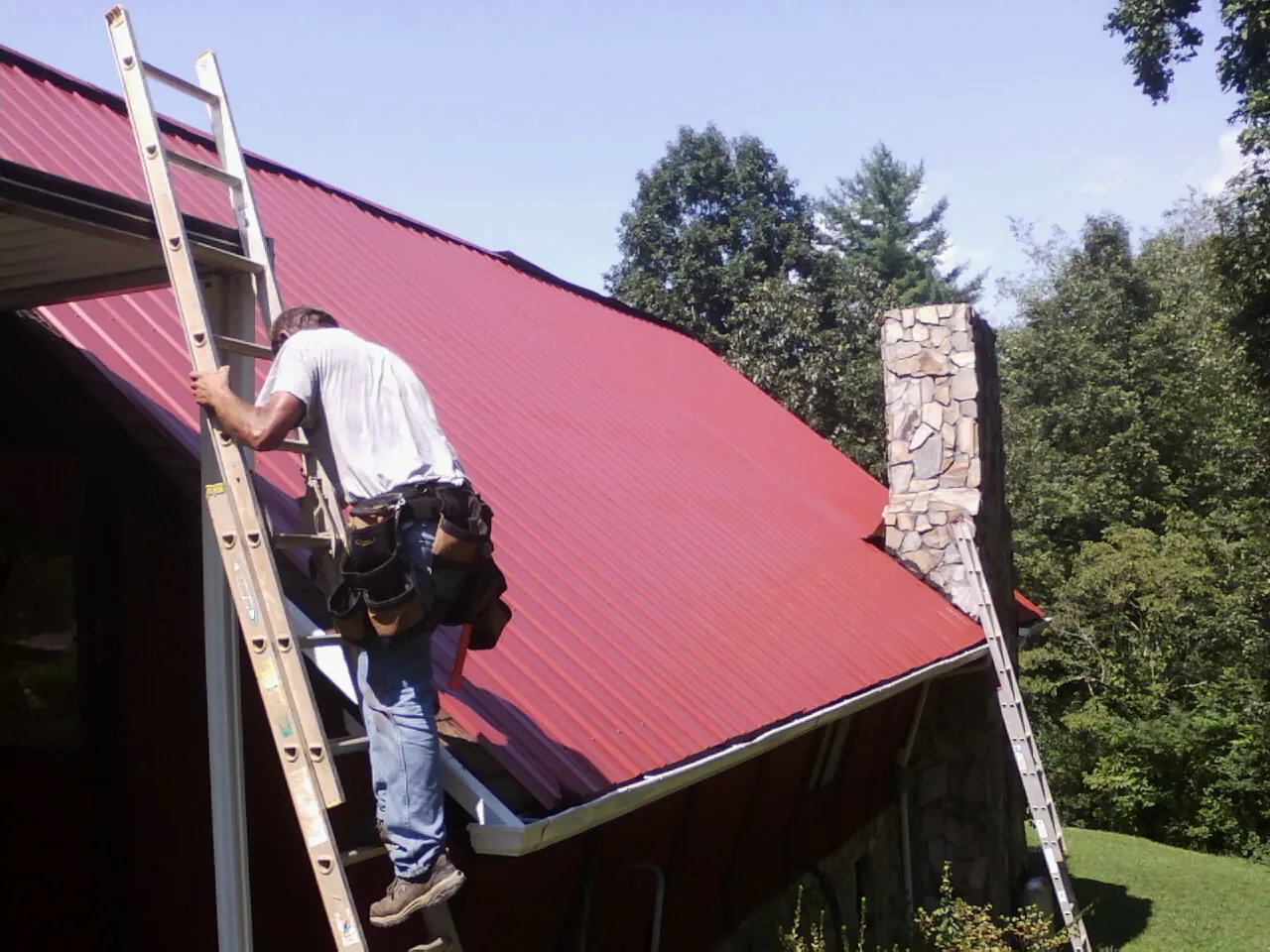 Roofer on a ladder beside a red metal roof and stone chimney — Soffit & Fascia Repair by Roofing Brothers in Suitland